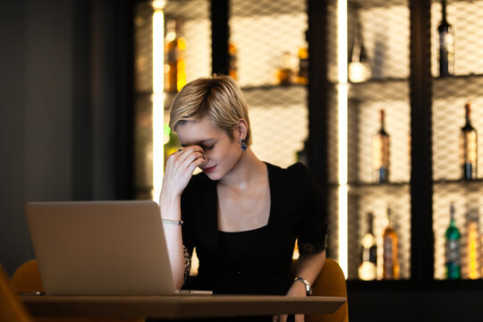 In a sophisticated bar, a young woman expresses fatigue or stress while working late on her laptop. The environment suggests an after-hours work scenario.