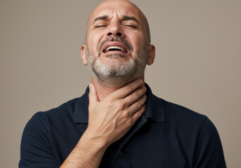 Portrait of a white man touching his neck and showing signs of discomfort or pain, suggesting sore throat, inflamed lymph nodes, or neck strain. Neutral gray background. Health and illness theme.