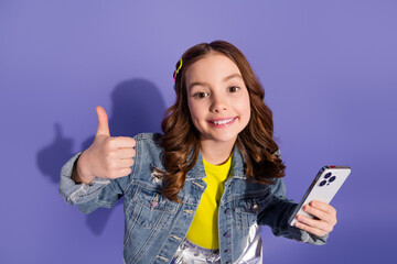 Happy young girl with long brown hair giving a thumbs up and holding a smartphone against a vibrant violet background