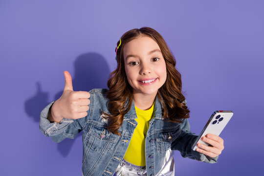 Happy young girl with long brown hair giving a thumbs up and holding a smartphone against a vibrant violet background