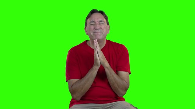 Man in red shirt desperately prays with hands clasped and eyes closed, expressing deep emotion and pleading, set against a green screen background for easy editing.  	