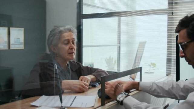 Cautious elderly woman making credit card payment and asking bank teller questions across service window