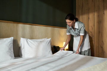 Housekeeping woman making bed with white sheets in a neatly arranged hotel room illuminated with...