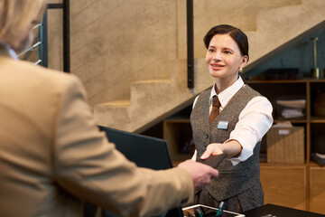 Smiling receptionist greeting customer at desk in modern office lobby, engaging in friendly interaction while assisting. Professional environment with shelves and staircase in background © pressmaster