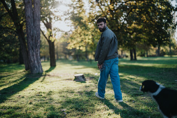 Fototapeta premium A man in jeans and jacket strolls through a lush green park, accompanied by his dog. The sunlight filters through the trees, creating a peaceful and relaxing atmosphere.