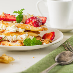 Belgian waffles with cottage cheese cream, strawberries, walnuts and mint on a white plate on a light table with a cup of coffee. Delicious, Sunday breakfast.
