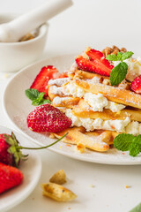 Belgian waffles with cottage cheese cream, strawberries, walnuts and mint on a white plate on a light table with a cup of coffee. Delicious, Sunday breakfast.