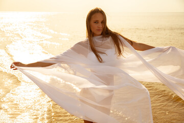 Beautiful Woman in White translucent Chiffon Dress flowing on Wind on the Beach at Sunset Sunshine.  Carefree Girl enjoying Sun at Seaside. Summer Vacation and Holidays. Freedom and Relaxation