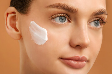 Hydration and skincare concept. Beautiful lady applying cream on her cheek, posing over beige background, closeup shot