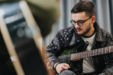 A young man wearing glasses practices guitar in a studio. The scene captures concentration,...
