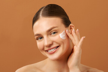 Fototapeta na wymiar Facial skincare. Lady applying moisturizing cream on cheek, caring for face and smiling, posing over beige studio background