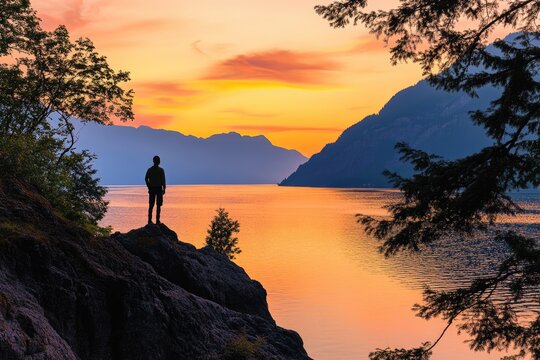 A lone figure gazes at the serene sunset over a lake, surrounded by majestic mountains and framed by trees. - Powered by Adobe
