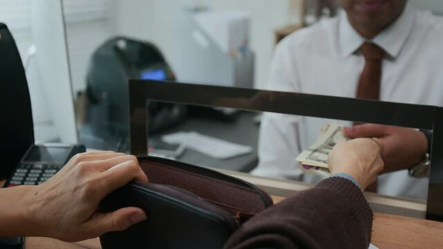 Close-up shot of unrecognizable elderly woman giving stack of dollar bills to male bank worker through glass teller window