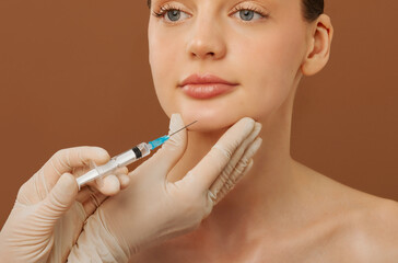 Beautiful young lady getting facial injection in chin, cosmetologist wearing gloves, holding...