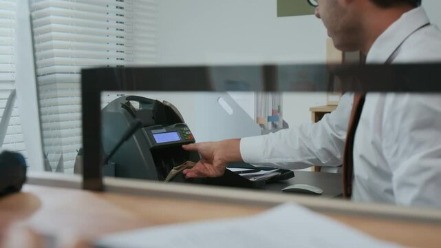 Close-up of man in white shirt and tie operating money counting machine behind glass teller window in bank office