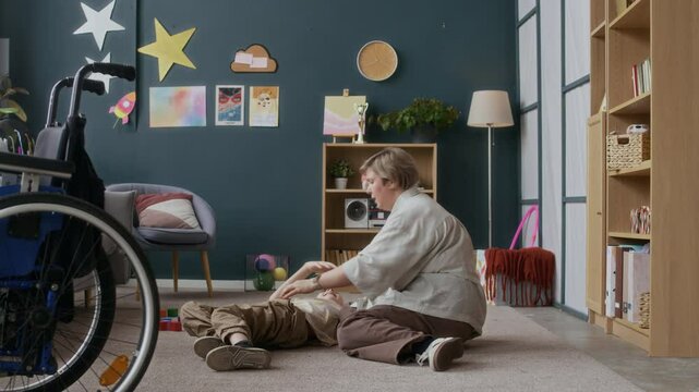 Long shot of caring Caucasian mom doing massage to teenage son with disability resting on floor in living room, mobilizing muscles strength