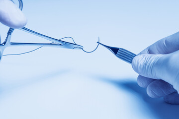 Operator hands holding a surgical needle threaded with non-absorbable suture. Sharp point and fine thread against a white background.