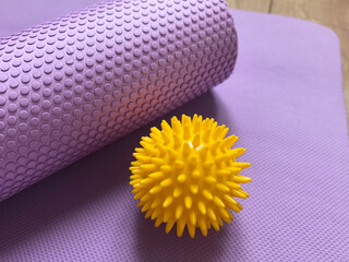 Sports equipment shown close-up: yellow massage ball and purple myofascial release roller behind it against the background of yoga, pilates and fitness mat lying on wooden floor. Fitness and health