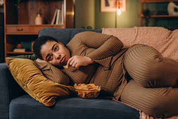 Obraz premium African American woman eats spicy chips lying on sofa in living room