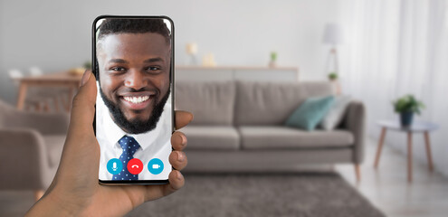 Naklejka premium A person is holding a smartphone displaying a smiling individual during a video call. The background reveals a cozy living room with a couch and plants, creating a warm atmosphere.