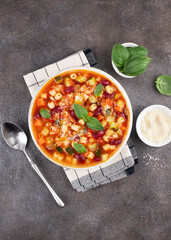 Minestrone soup with fresh vegetables, beans, pasta, parmesan cheese and basil on a gray background. Italian cuisine.