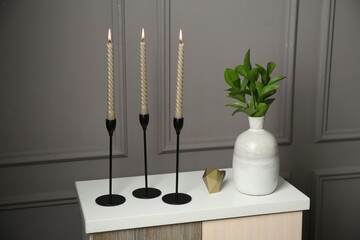 Elegant candlesticks with burning candles, branches in vase and other decor on dresser near grey...