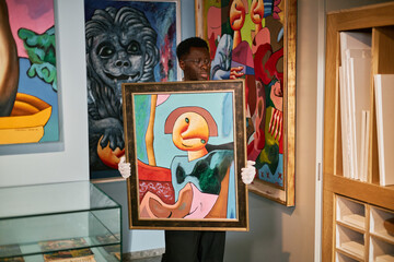 Art gallery staff holding abstract painting while showcasing various vibrant artworks hanging on...