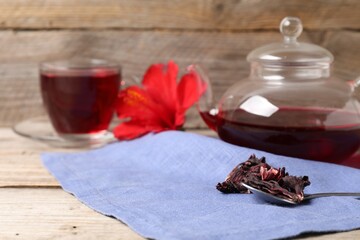 Delicious hibiscus tea in glass cup, teapot, dry roselle sepals and spoon on wooden table, closeup. Space for text