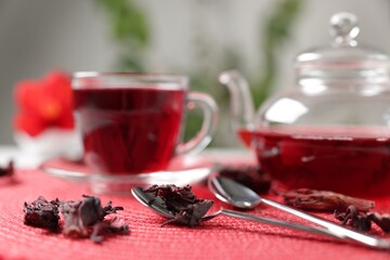 Delicious hibiscus tea in glass cup, teapot, dry roselle sepals and spoons on red mat, closeup
