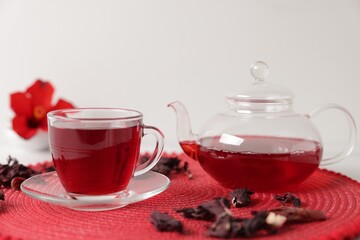 Delicious hibiscus tea in glass cup, teapot and dry roselle sepals on red mat, closeup