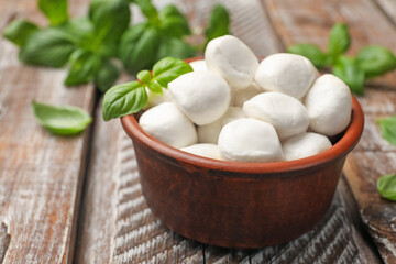 Tasty mozzarella cheese balls in bowl and basil on wooden table, closeup