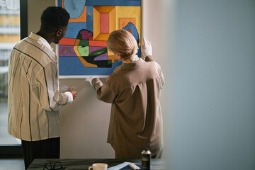 Two people engaged in closely examining vibrant modern art piece in gallery setting. Artwork featuring geometric shapes on vibrant colored background