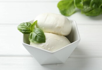Pieces of delicious mozzarella cheese and basil in bowl on white wooden table, closeup