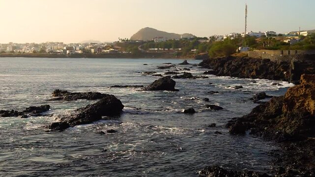 Sunset view over the rocky coastline of Palmarejo district in Praia, Cape Verde, with urban buildings and hills in the background.
