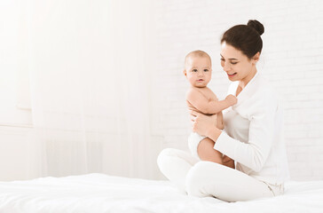 Happy maternity. Young mother sitting with baby on bed, looking at child with love and care, copy space