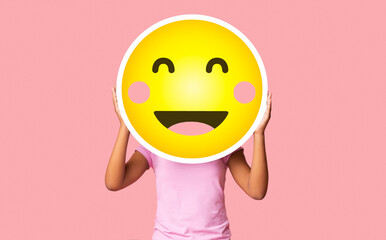 Black girl hiding face behind happy shy emoji emoticon, standing over pink studio background with...