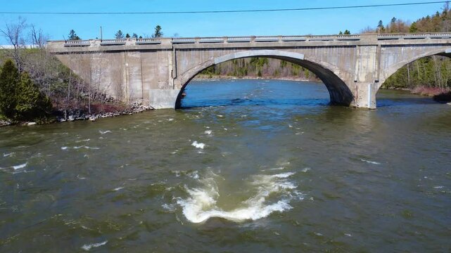 A rock creates eddies on the surface of a flooded river passing under the arch of an old concrete bridge. Mitis River, Arthur-Bergeron Bridge, Quebec, Canada, 2025.