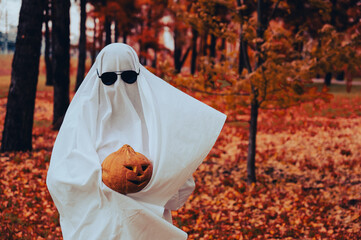 Woman in white sheet holding jack o'lantern against autumn forest background. Halloween costume. 
