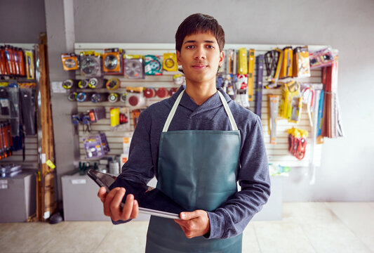 Young latin american shop assistant wearing apron holding digital tablet in hardware store