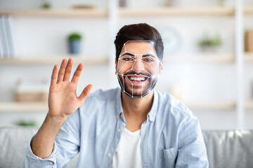 Obraz premium Indian man is using facial recognition technology in a home setting. He is smiling while waving, with a digital overlay highlighting his facial features for identification.