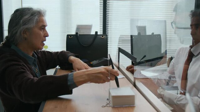 Male bank employee showing elderly woman where to sign her documents across glass teller window in office