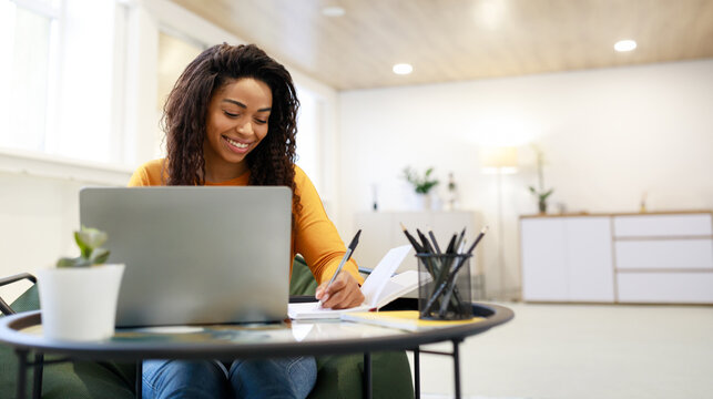 Smiling young black woman sitting at desk working on laptop taking notes in notebook, happy millennial female study watching webinar using computer writing check list, banner panorama, free copy space