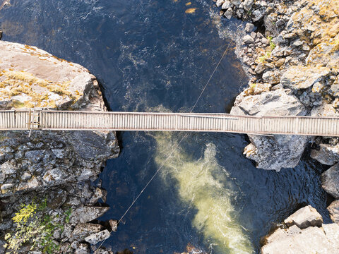 Aerial drone view: a narrow wooden suspension bridge crosses a turbulent river in a rocky canyon of Norway's wild nature.