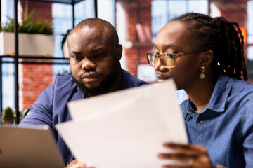 Couple filling in spreadsheets on laptop by entering data from report papers, calculating totals and analyzing figures at the end of the month. African american people working from home.