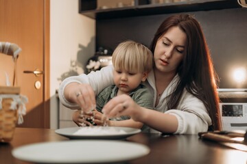 Focused toddler explores flour on a plate while holding a spoon next to his supportive mom a touching everyday scene showing authentic learning playful growth and mindful connection in family life