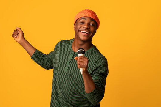 Cheerful Black Millennial Guy Holding Microphone And Singing At Camera, Young Joyful African American Hipster Man Enjoying Karaoke, Having Fun While Standing Over Yellow Studio Background, Copy Space