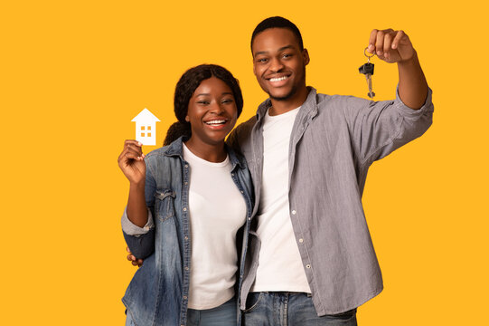 New Home Owners Concept. Happy african american couple holding paper house and keys, smiling young black spouses celebrating buying property, standing isolated on yellow background, copy space