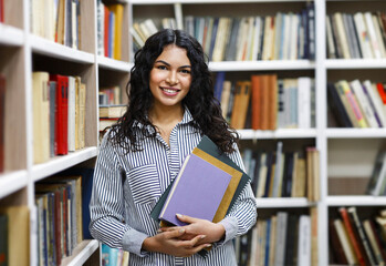 Academic Concept. Happy smiling latina woman holding workbooks leaning on bookshelves in campus...