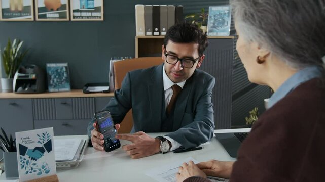 Over the shoulder shot of Indian bank employee in suit answering questions of elderly woman about smartphone app in bank office