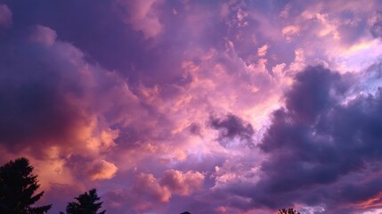 A breathtaking sky filled with shades of purple and orange clouds creating a vibrant and dynamic sunset over the distant silhouette of trees.